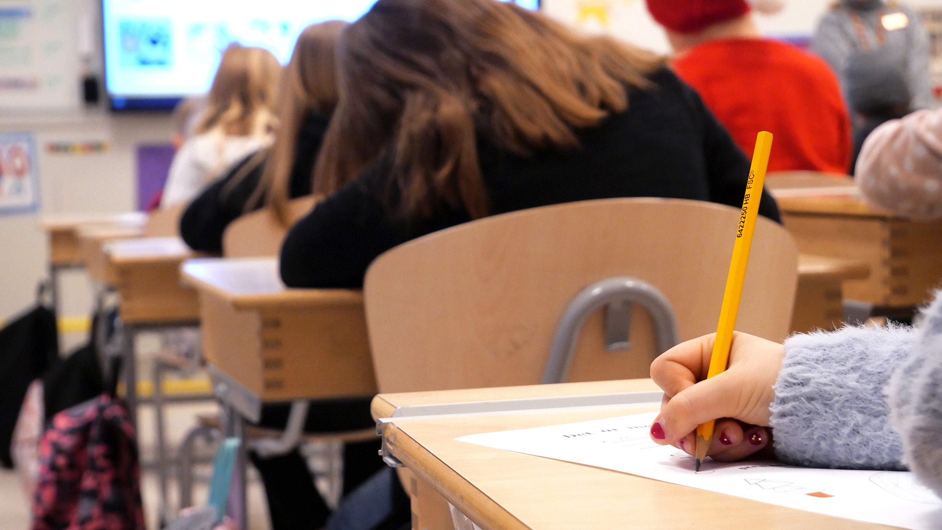 Bilden är tagen i ett klassrum och är en närbild på en hand som skriver med en blyertspenna. På bilden syns även ryggtavlor på fyra andra elever. 