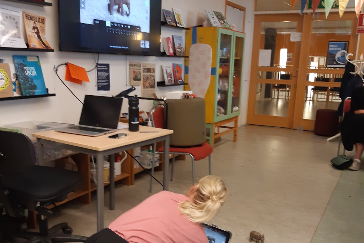 Bilden visar en person som sitter på golvet med en bärbar dator framför sig, framför datorn står en leksaks elefant. Bredvid personen är en vägg och på väggen sitter en bildskärm som visar leksaks elefanten som står på golvet. 