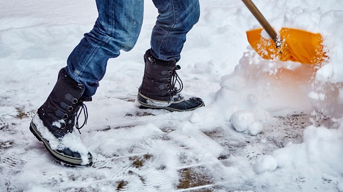 Bilden visar hur någon i jeans och vinterkängor skyfflar snö från en infart till en fastighet.