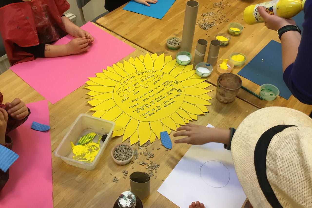 Bilden visar barn kring ett bord  som provar tryckteknik med disktrasa och toarullar. Dem trycker blomblad, pickar färg och klistrar fast solrosfrön på färgglada pappersark. 