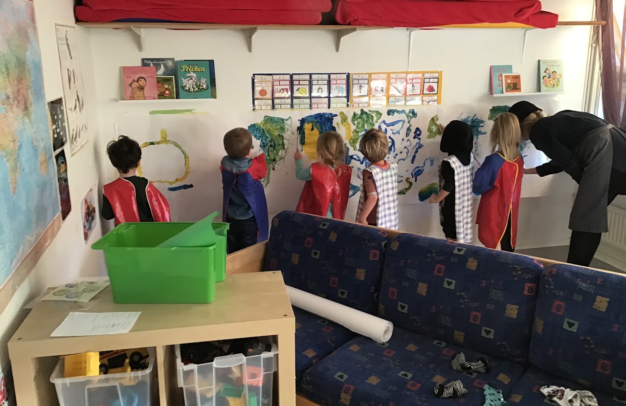 Bilden visar sex stycken barn och en lärare som står och målar på ett stort papper som är uppsatt  längst med en vägg. 