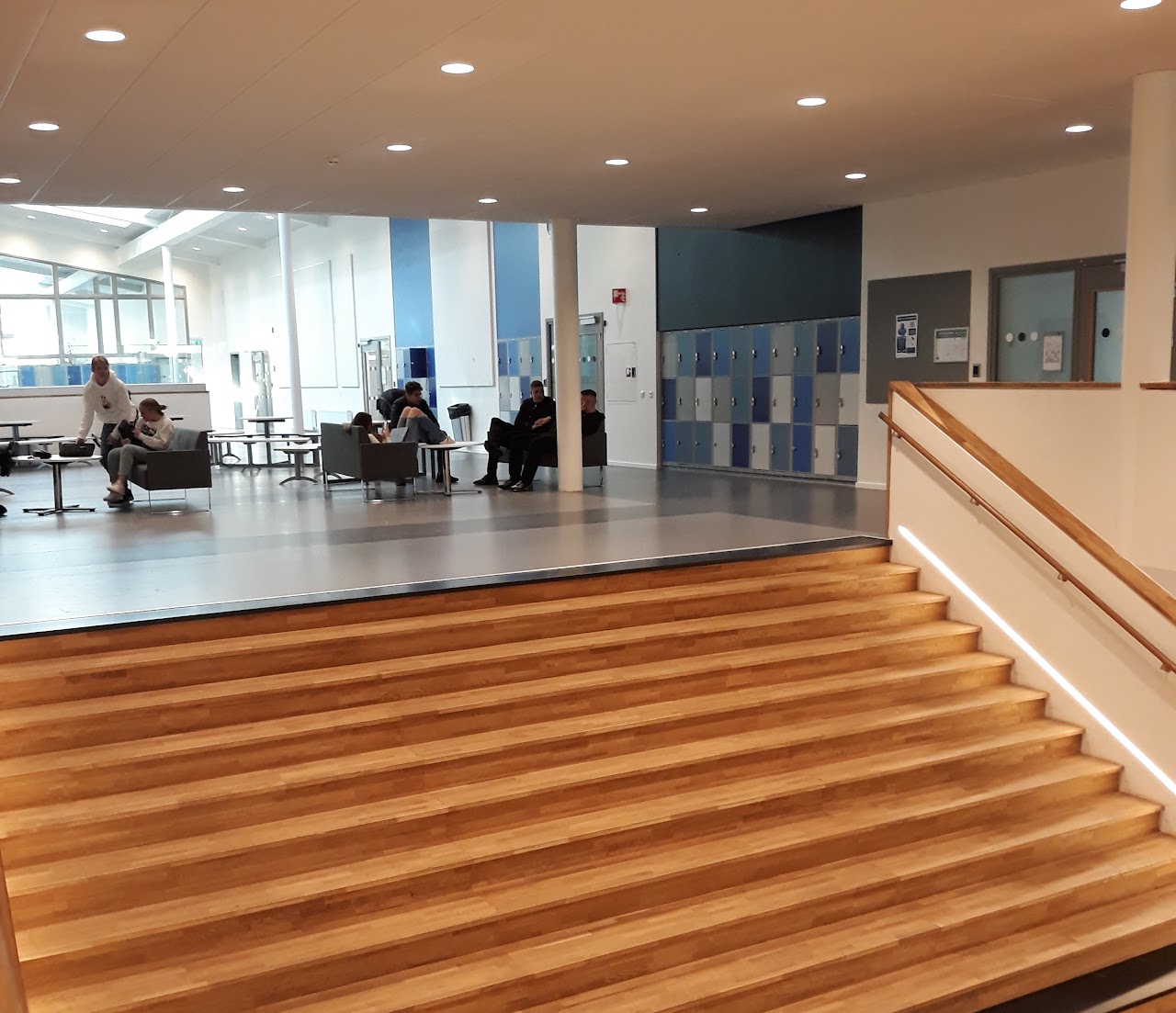 Bilden visar övre delen på Stadsparkskolan trappa, ovanför trappan är ett uppehållsrum med skåp längst väggarna. Det står soffor, fåtöljer och bord och på några ställen sitter elever. 