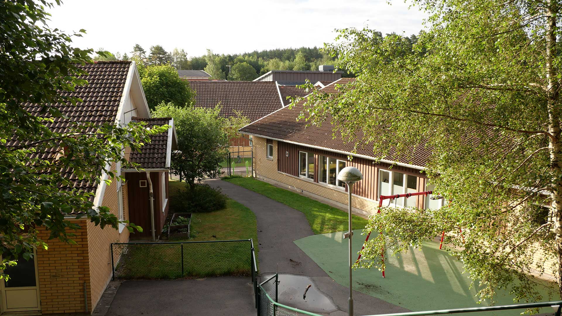 Skolan består av gula tegelbyggnader i ett plan. Mellan  de tvp byggnaderna på bilden , syns en 
inhägnad skolgård. 