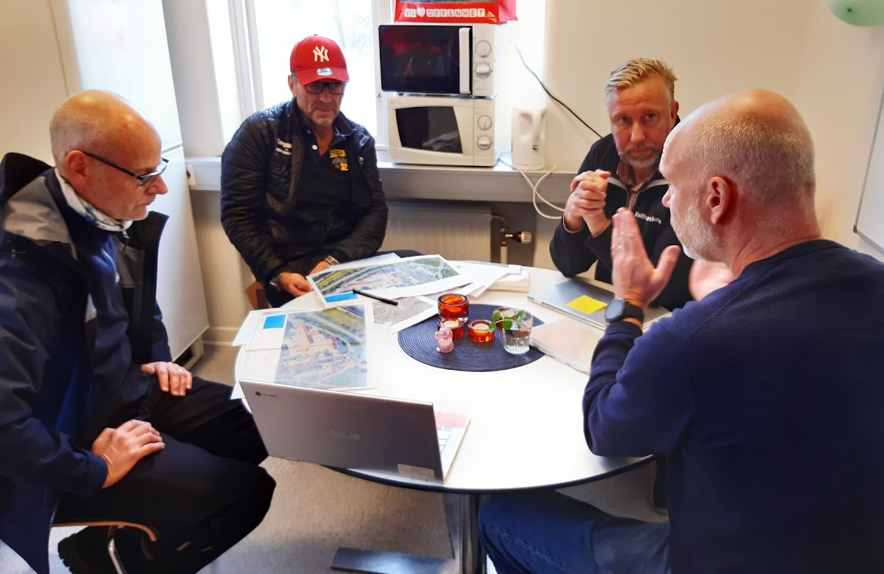 Bilden visar fyra män som sitter runt ett bord med kartor och ritningar. 