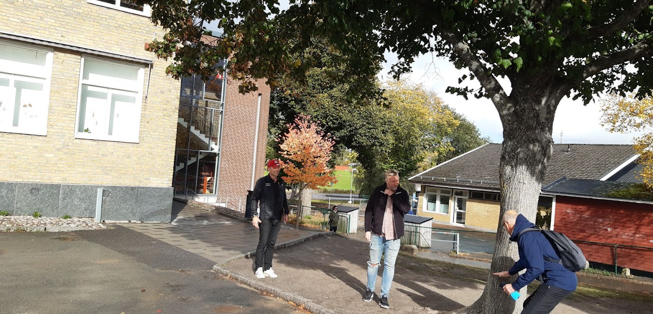 Bilden visar tre män som står under ett träd på en skolgård, en av dem måttar med ena handen på trädstammen. 