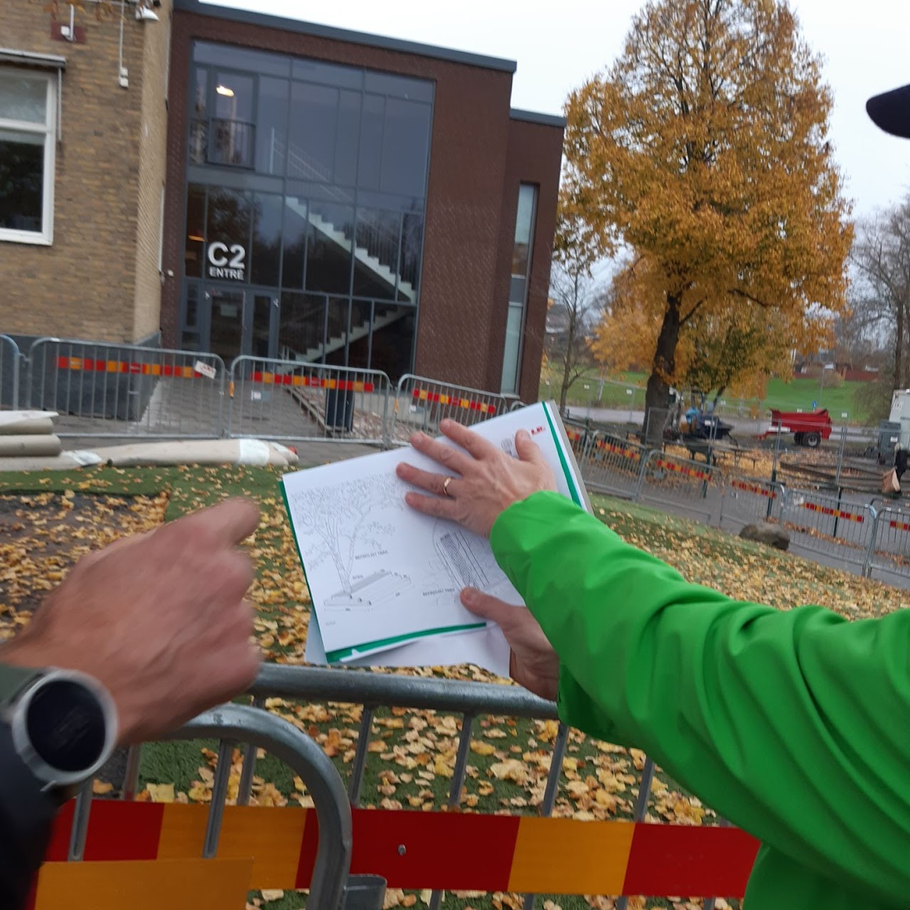 Bilden visar en arm som håller upp en planritning framför ett avspärrat område utanför en skolentré.
