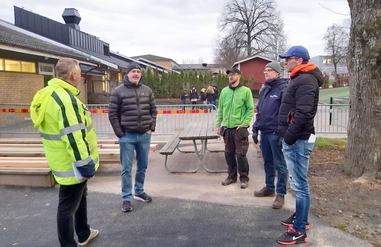 Bilden visar fem män som står framför en avspärrning och en hög med byggmaterial på en skolgård. 