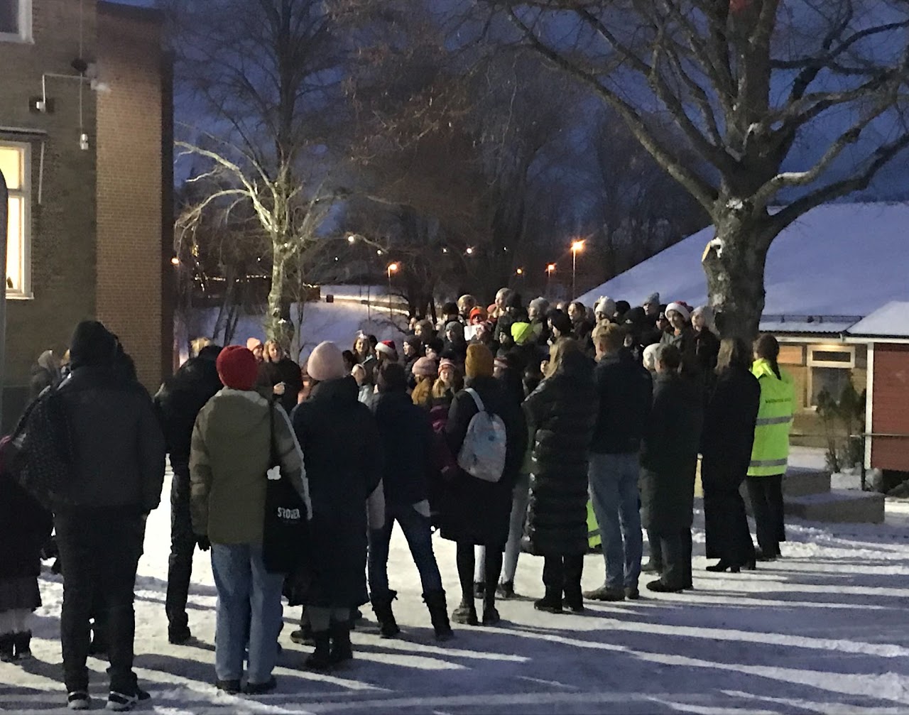 Bilden visar en större grupp människor i vinterkläder, en kväll på en upplyst vintrig skolgård.