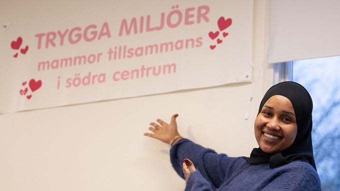 Bilden visar en leende person med lila stickad tröja och svart slöja. I bakgrunden syns en skylt med texten: Trygga miljöer – Mammor tillsammans i södra centrum.