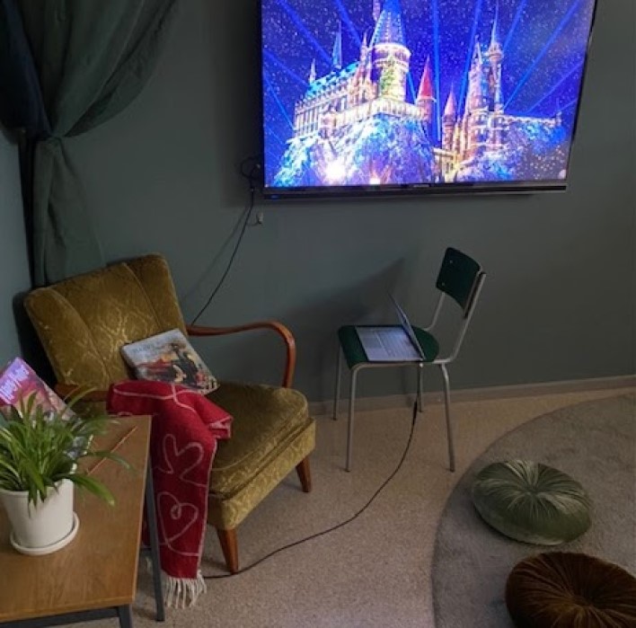 Bilden visar ett hörn på ett rum där det står en grön fåtölj med en röd pläd över det ena armstödet och en bok som står på sitsen lutad mot ryggstödet. Bredvid fåtöljen står ett litet bord med ytterligare en bok på samt en kruka med en grön växt. På väggen hänger en bildskärm som visar ett upplyst slott. Nedanför bildskärmen står en stol med en bärbar dator på. På golvet framför fåtöljen ligger sittpuffar i olika färger. 