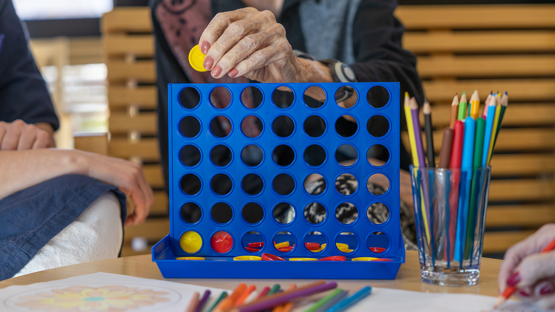 Bild på äldre person som spelar sällskapsspel vid ett bord omgiven av två andra personer. I fokus på bilden är den äldre personens hand.