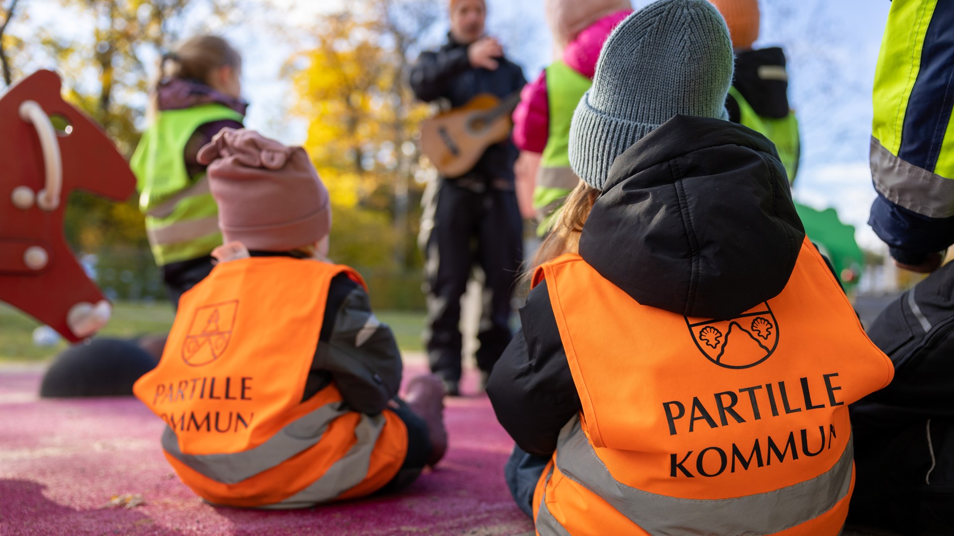 Bild som visar förskolebarn som sitter på marken och lyssnar på en pedagog som spelar musik.