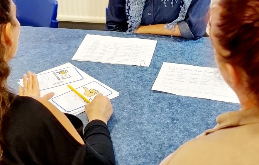 Bilden visar tre personer som sitter vid ett bord, på bordet ligger schema och bildstöd. 