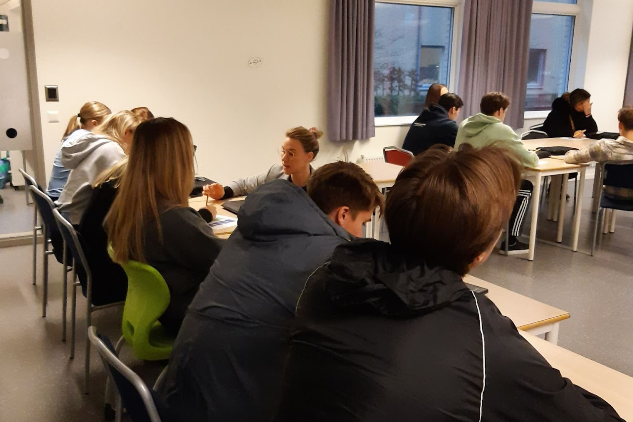 Bild på en rad med skolbänkar där det sitter flera elever, en pedagog sitter på framför en av bänkarna och hjälper en elev med en uppgift. 