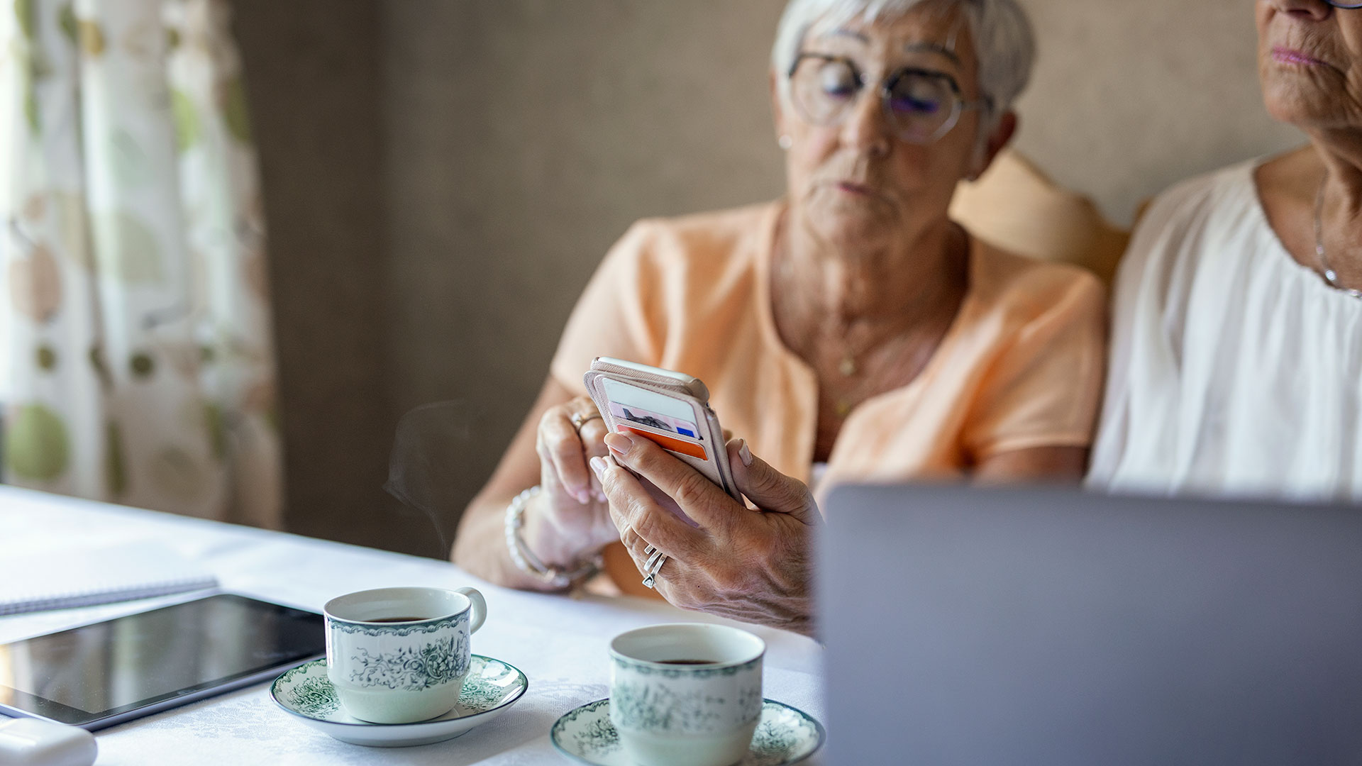 Bilden visar tv&aring; personer som sitter vid ett bord och tittar p&aring; en mobiltelefon. P&aring; bordet finns &auml;ven en uppslagen dator, en l&auml;splatta och ask med h&ouml;rlurar samt tv&aring; kaffekoppar.