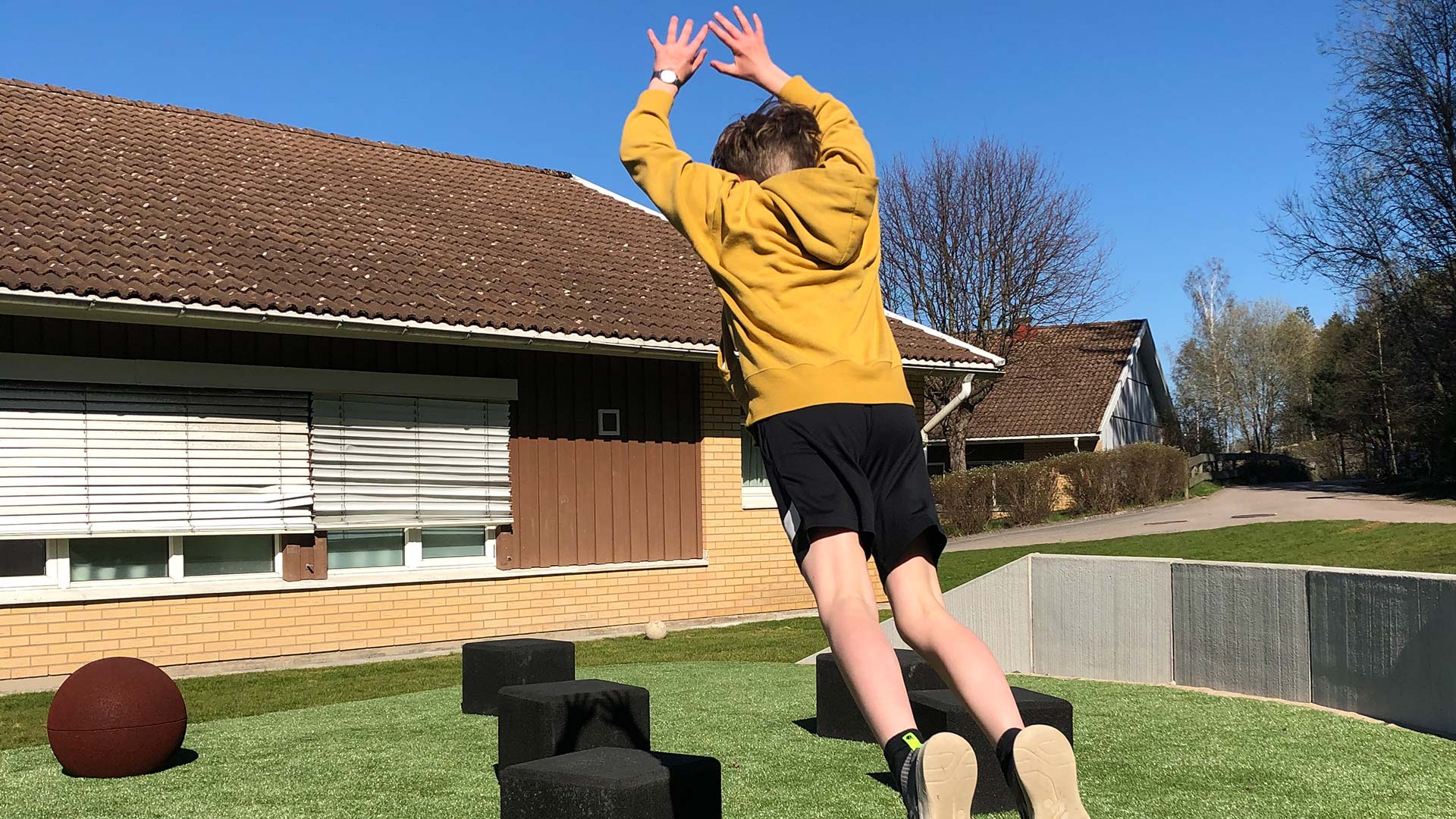 En ung kille iförd svarta shorts och gul tröja med huva, hoppar jämfota från en sten på skolgården och stäcker armarna upp i luften under hoppet. 