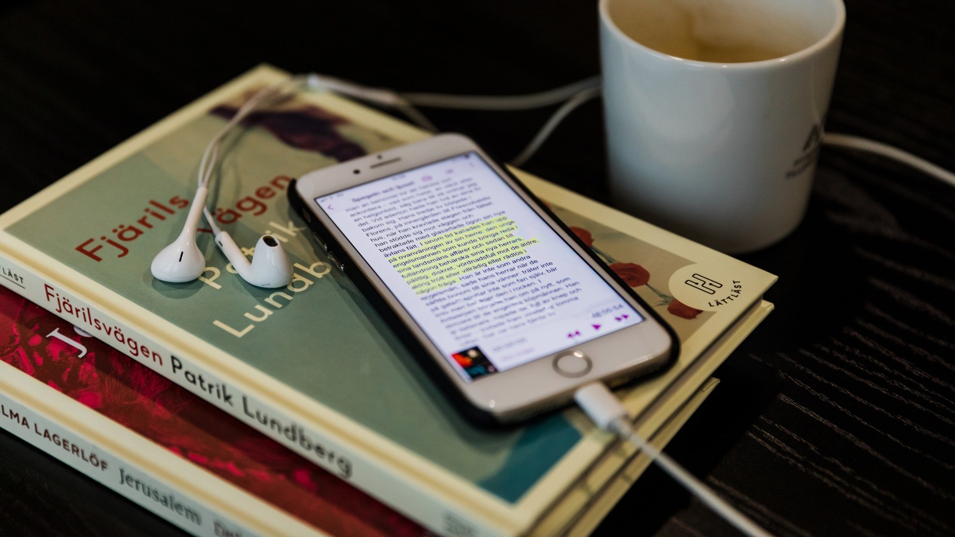 En telefon ligger ovanpå ett par böcker på ett bord. Bredvid står en kaffekopp. Skärmen på telefonen visar en boksida från Legimusappen, en del av texten är markerad i gult. Bilden är tagen snett från sidan.