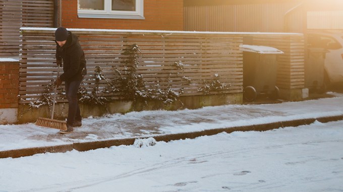 Bilden visar en vinterklädd person som sopar bort snö från trottoar utanför ett bostadshus.