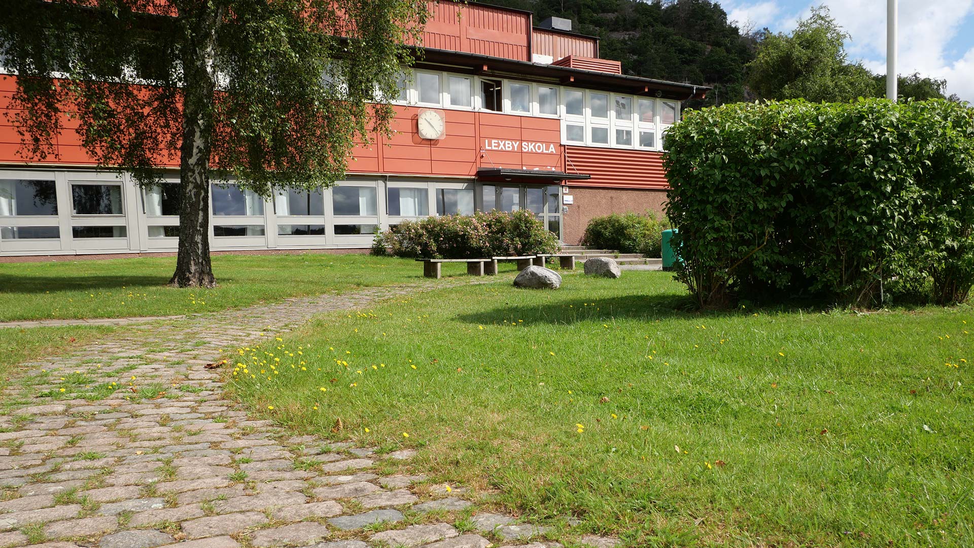 Bilden visar Lexby skola, en rödfärgad byggnad i två vångar. Framför skolan syns en björk och några buskar, en stenlagd gång, några sittbänkar samt en flaggstång. 