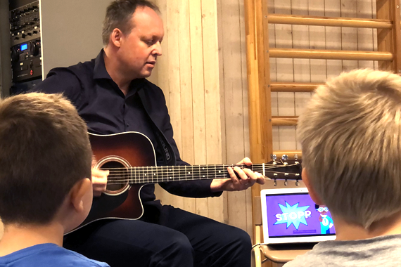 Bilden visar musikläraren Magnus Jonsson som sitter med en gitarr i knät, runt honom i en ring sitter elever.  