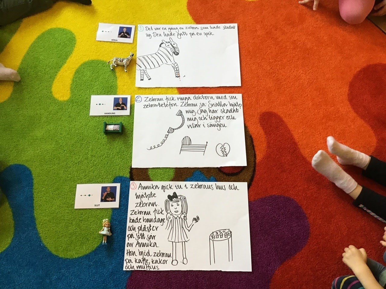 Bilden visar en kort saga på tre ark. Bredvid vardera ark ligger ett tecken-kort samt en plastfigur som motsvarar den illustrerade bilden på vardera ark. Sagan i sin helhet ligger på en regnbågsfärgad matta och några barnfötter syns i kant. 