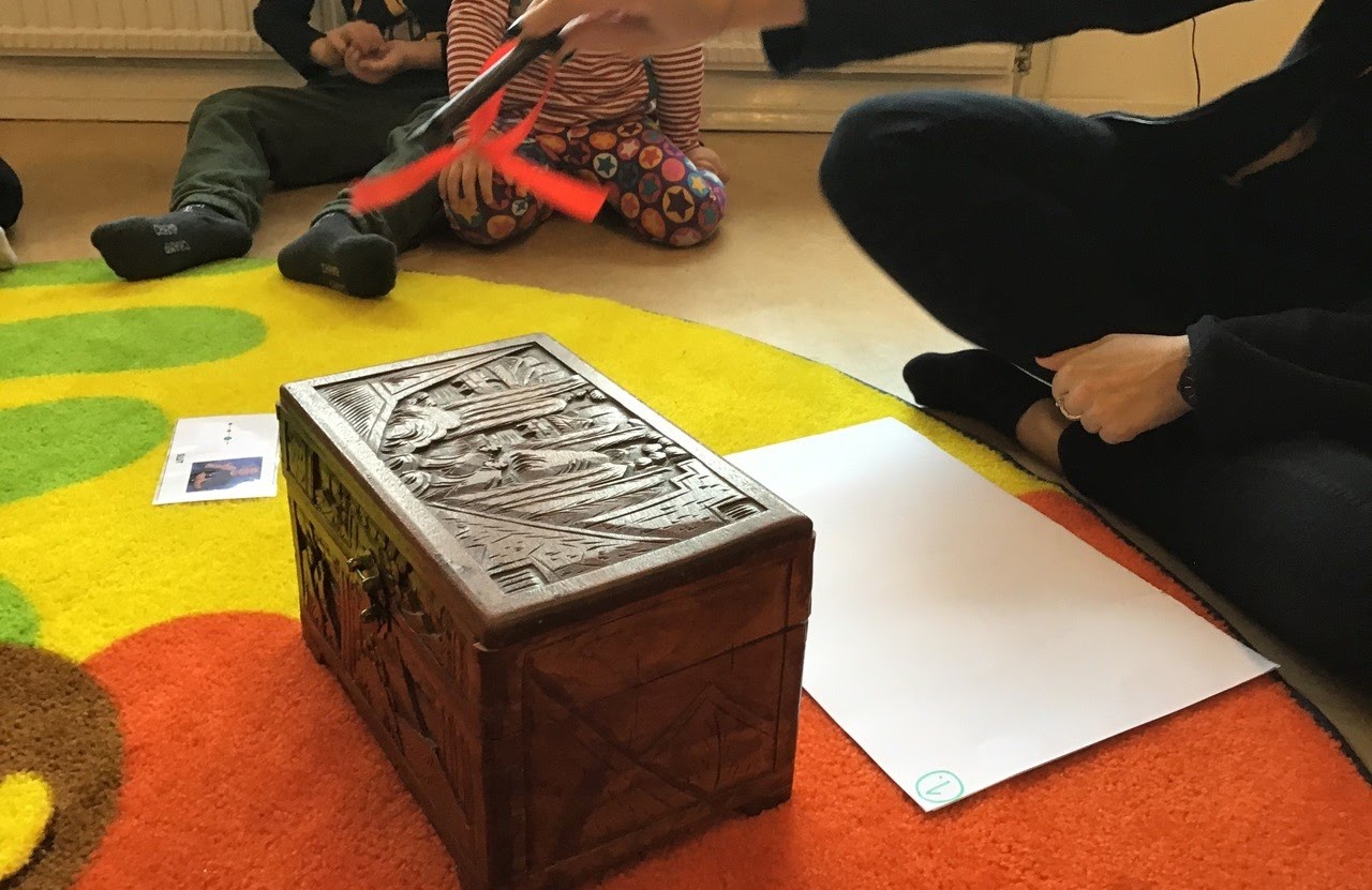 Bilden visar en mörkbrun kista i trä som står på en färgglad matta, en pedagog sitter framför lådan och håller i ett spö ovanför lådan. Bredvid pedagogen sitter två barn. 