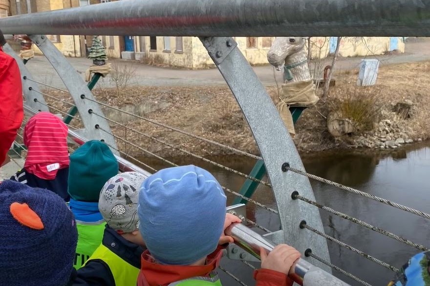 Bilden visar en grupp barn med mössor som håller sig i ett räcke på en bro som går över en å. På brostolparna sitter konstverk i form av hästhuvuden som barnen tittar på.  