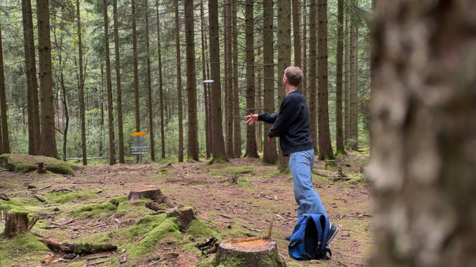 Bilden visar en person som kastar iväg en frisbee mot en discgolfkorg i skogsmiljö.