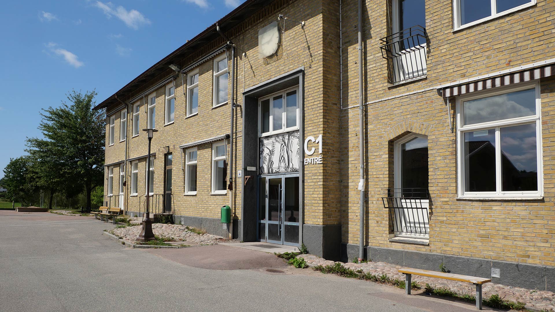 På bilden syns en tvåvåningar hög skolbyggnad i gult tegel. Framför skolbyggnaden finns skolgården med flera sittbänkar.