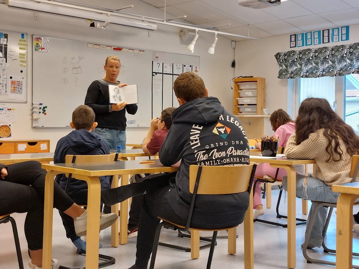 Bilden visar en lärare som står längst fram i ett klassrum och håller upp ett uppslag ur en bok, i klassrummet sitter flera elever och tittar på läraren. 