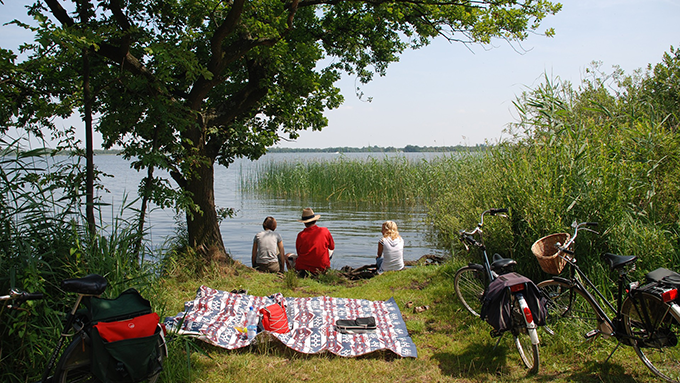 Tre personer sitter vid badsjö med en picknickduk intill. I förgrunden står tre cyklar parkerade.