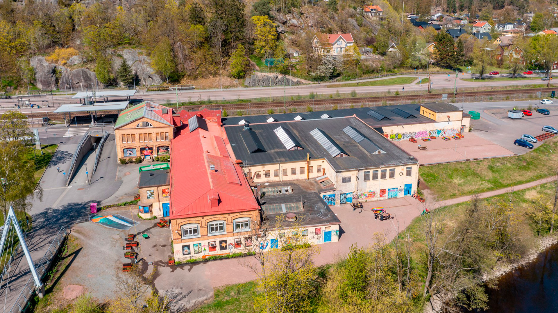 Bilden är tagen med drönare och visar verksamhetsbebyggelse i ett kvarter som ligger mellan ett vattendrag, bilväg och järnvägsräls.
