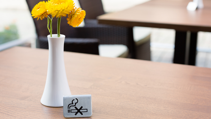 Bild på kafébord med vit vas med gula blommor i och en skylt med ett pictogram som visar att rökning är förbjudet.
