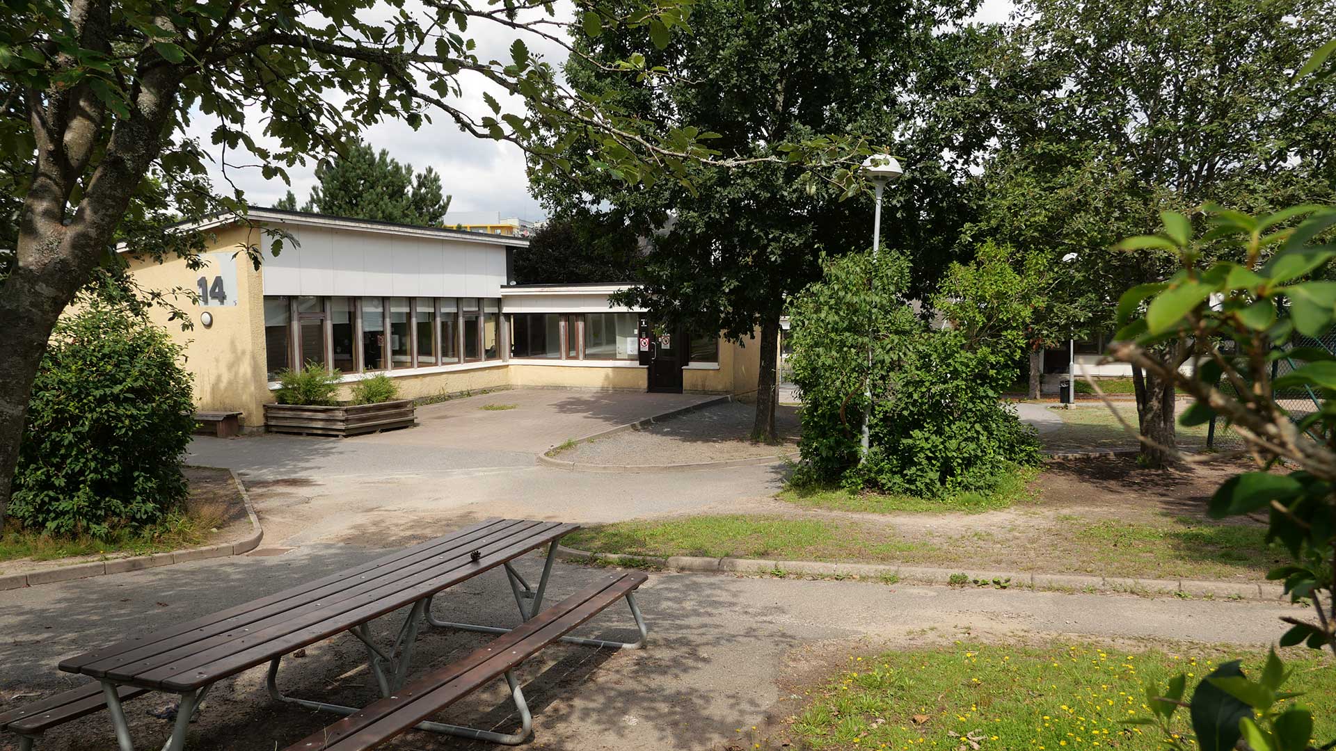 En gul enplansbyggnad i vinkel och med skolgård framför där det står en bänk och ett bord. 