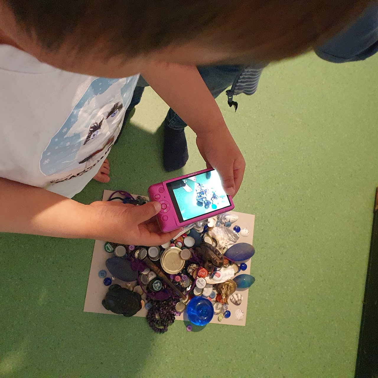 Bilden visar ett barn som håller i en rosa digitalkamera över ett papper där det ligger massor av kapsyler, stenar och glasbitar i olika färger. 