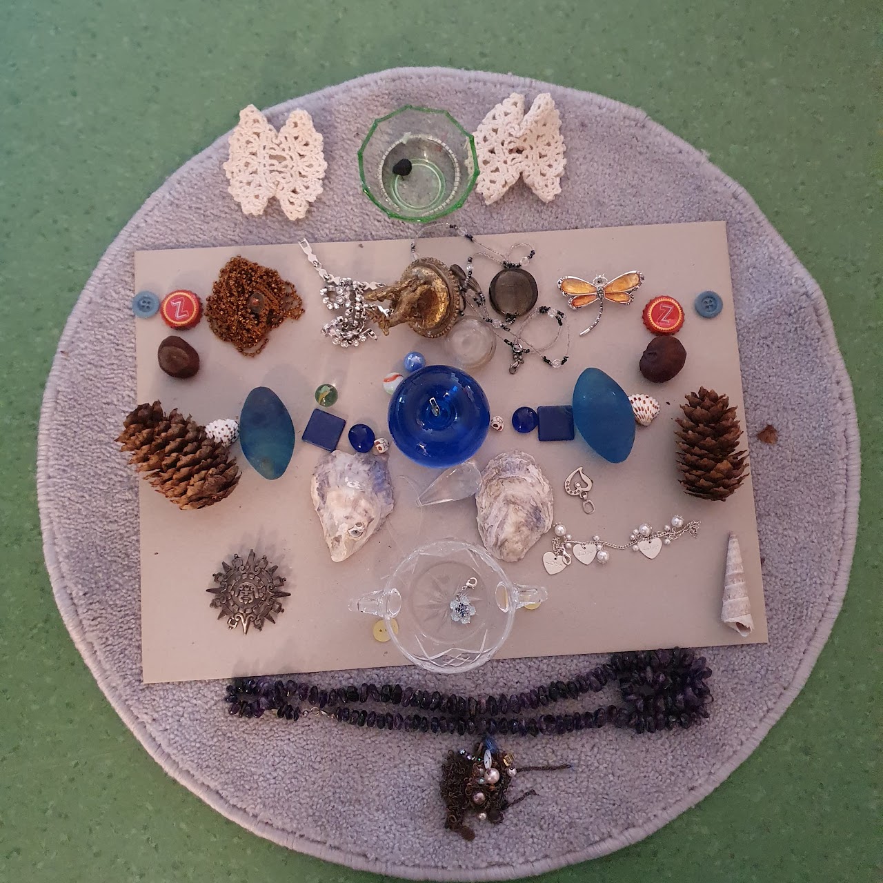 Bilden visar en rund grå matta med diverse föremål på så som kottar, glasbitar, piprensare och kapsyler. 