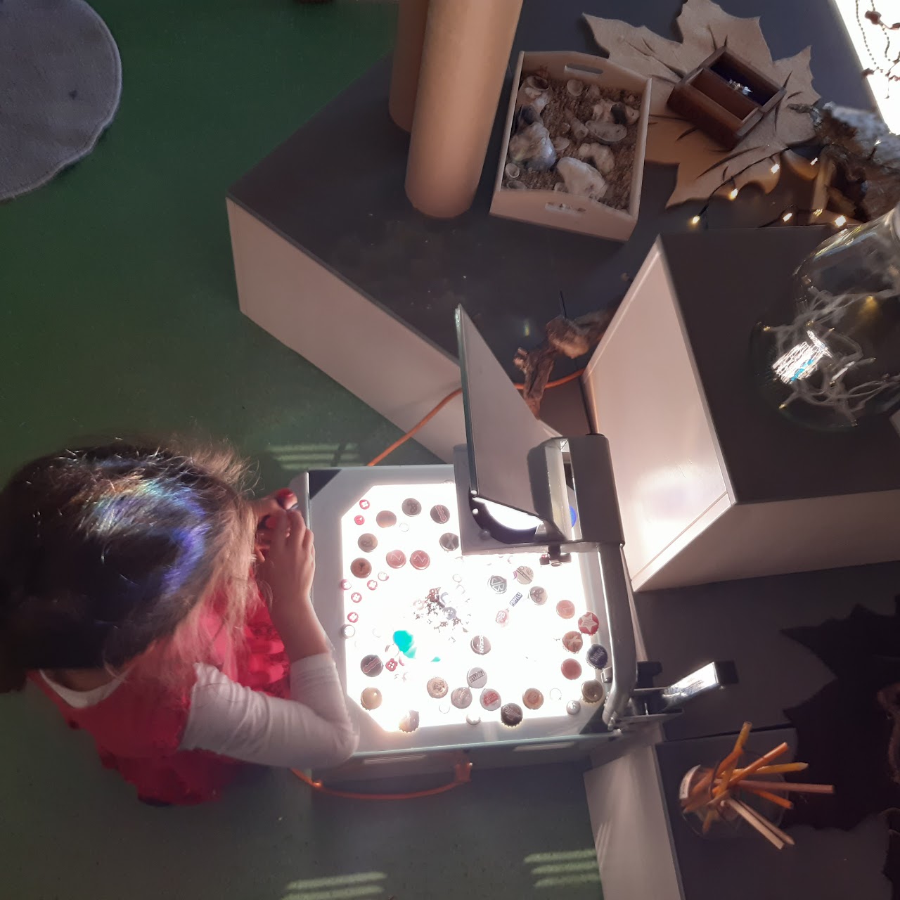Bilden visar ett barn som sitter framför en tänd overhead där det ligger knappar, kapsyler och pärlor. 