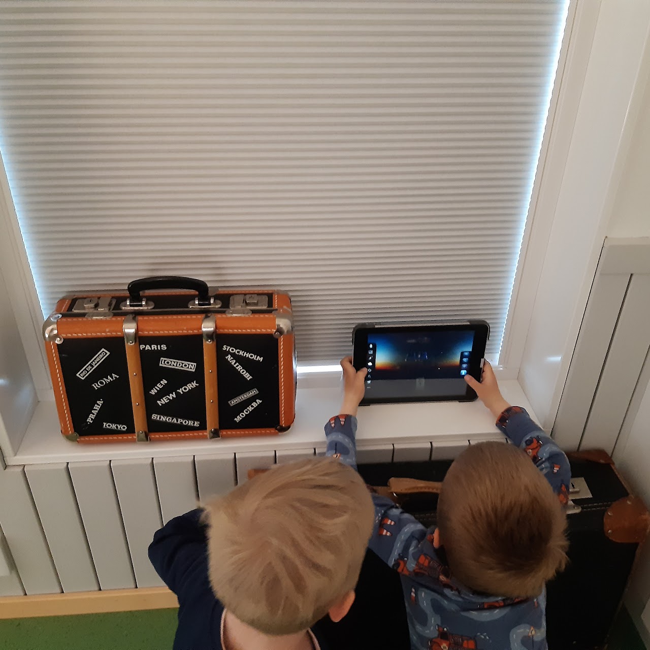 Bilden visar två barn som sitter på knä framför en fönstersmyg, det ena barnet håller en läsplatta i händerna som båda barnen tittar på. Bredvid läsplattan i fönstersmygen står en liten gammaldags resväska. 