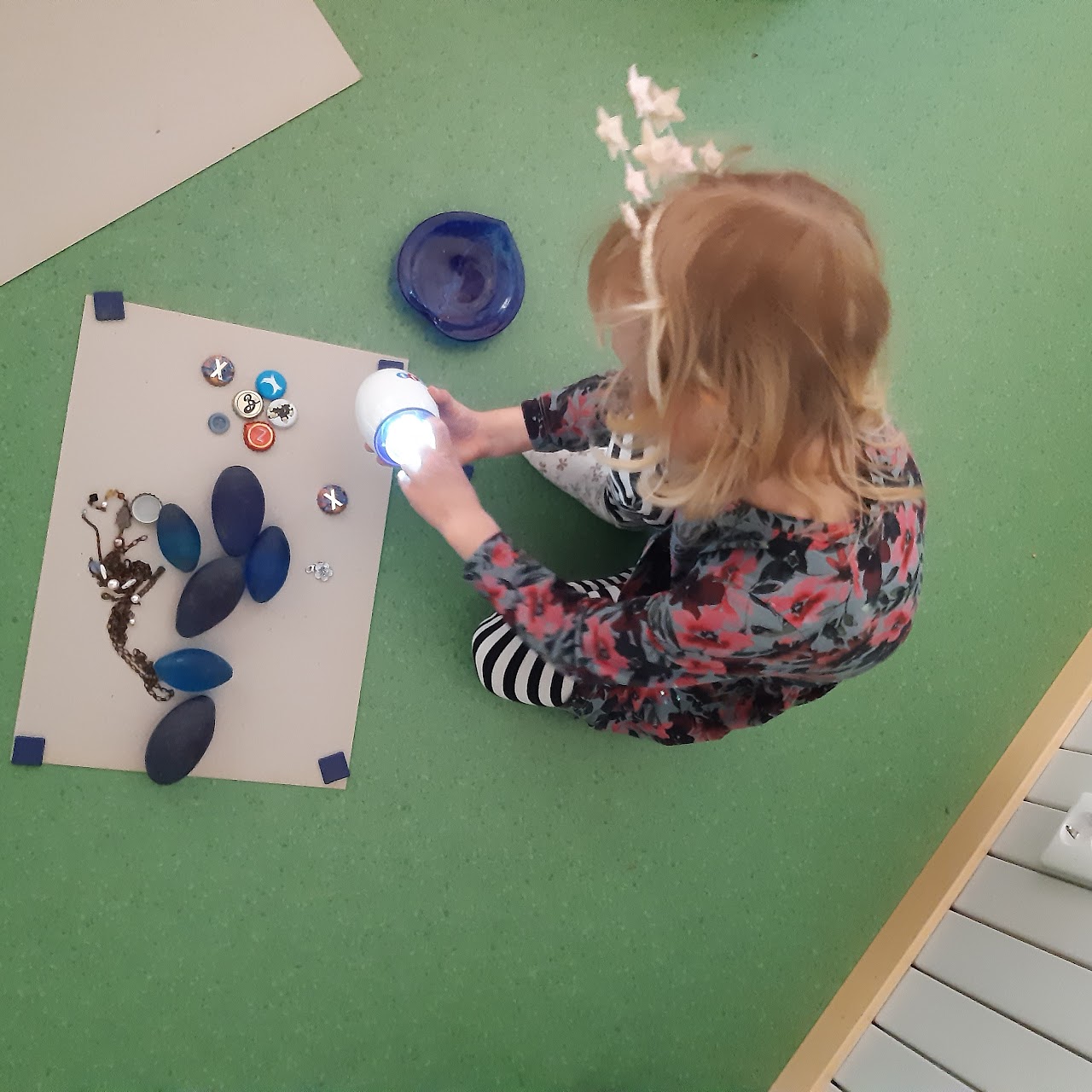 Bilden visar ett barn som sitter på ett grönt golv och håller i ett äggformat mikroskop som lyser. Framför barnet ligger ett papper på golvet och på pappret ligger stenar, knappar och pärlor i olika former och färger.  