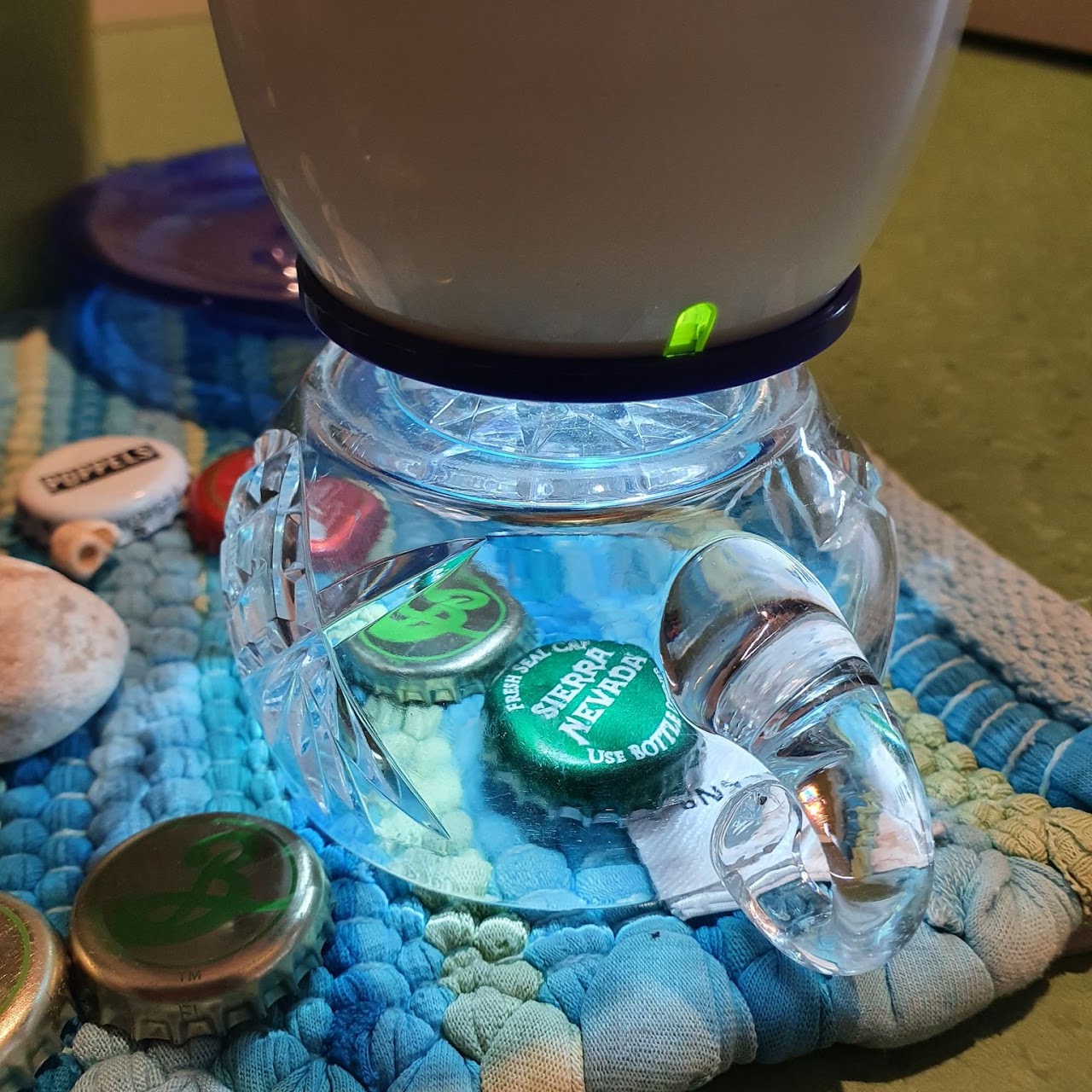 Bilden visar ett äggformat mikroskop som står på en kopp i glas. Under koppen ligger två stycken kapsyler på en trasmatta. 