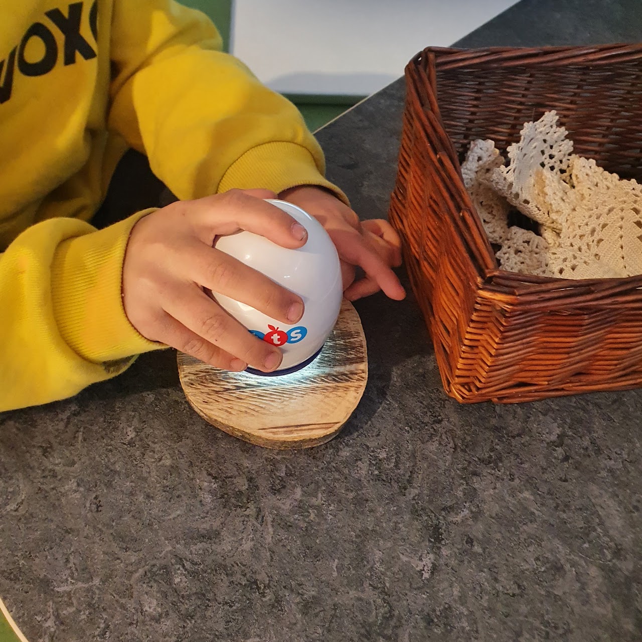 Bilden visar ett par barnhänder som håller i ett äggformat mikroskop mot en träbit, bredvid står en flätad korg med virkade dukar i.  