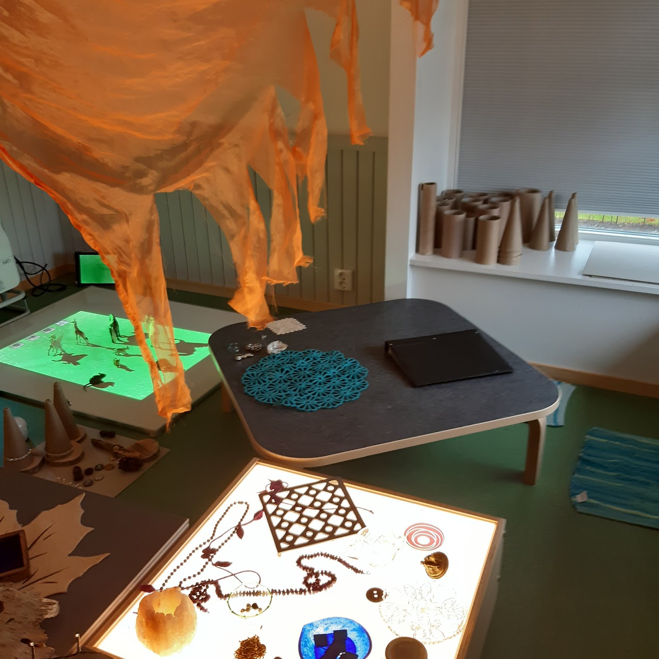 Bilden visar Tinker-rummet, där det står ett ljusbord med föremål på, ett mindre fyrkantigt bord står intill. Bortanför de lilla bordet ligger en Whiteboard på golvet där en grön bild projiceras. I taket hänger orange flik-klippt tyg och i fönstret står flertalet pappersrullar och papper koner. 
