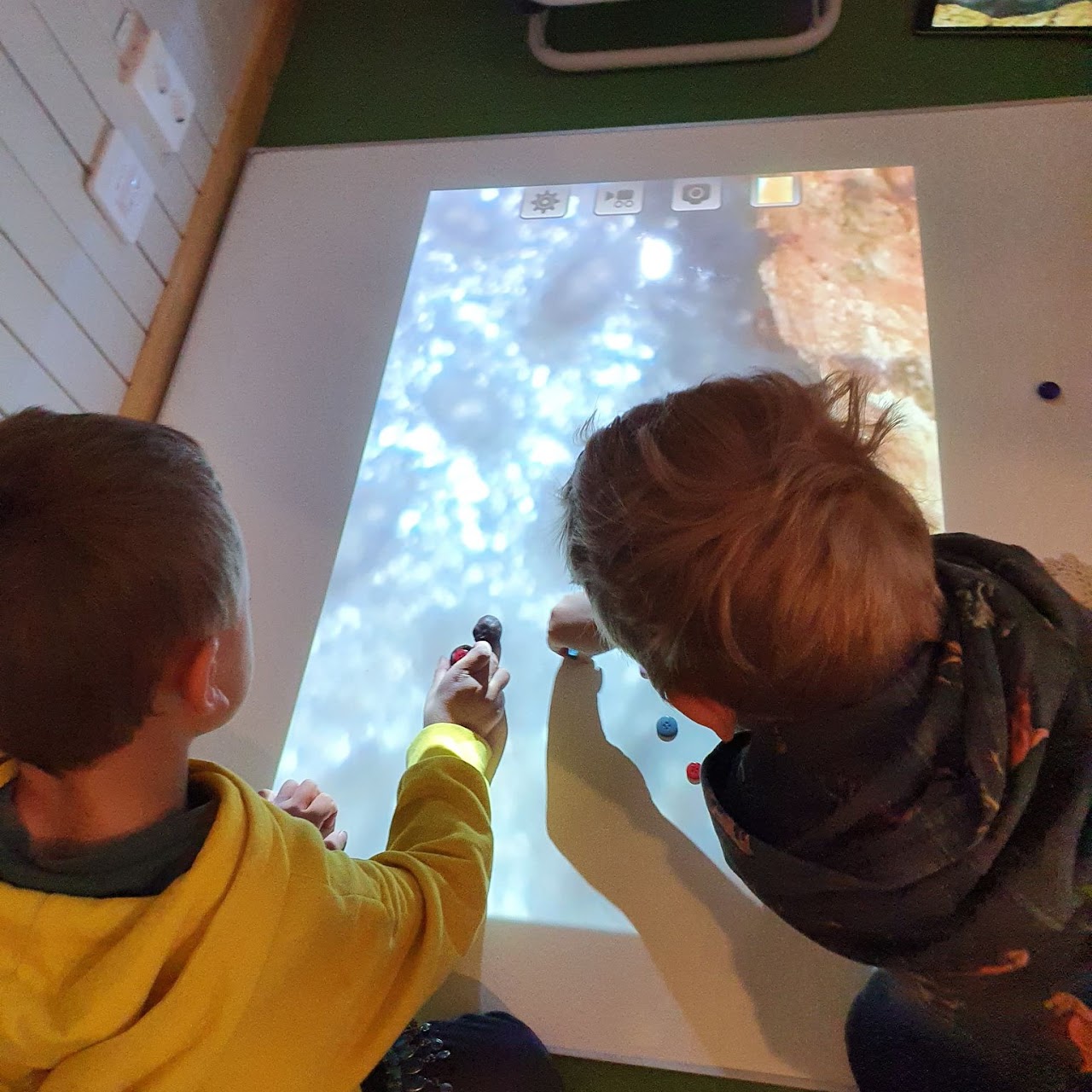 Bilden visar två barn som sitter på knä framför en Whiteboard som ligger på golvet. På Whiteboarden projiceras en bild och barnen har lagt ut knappar på bilden. 