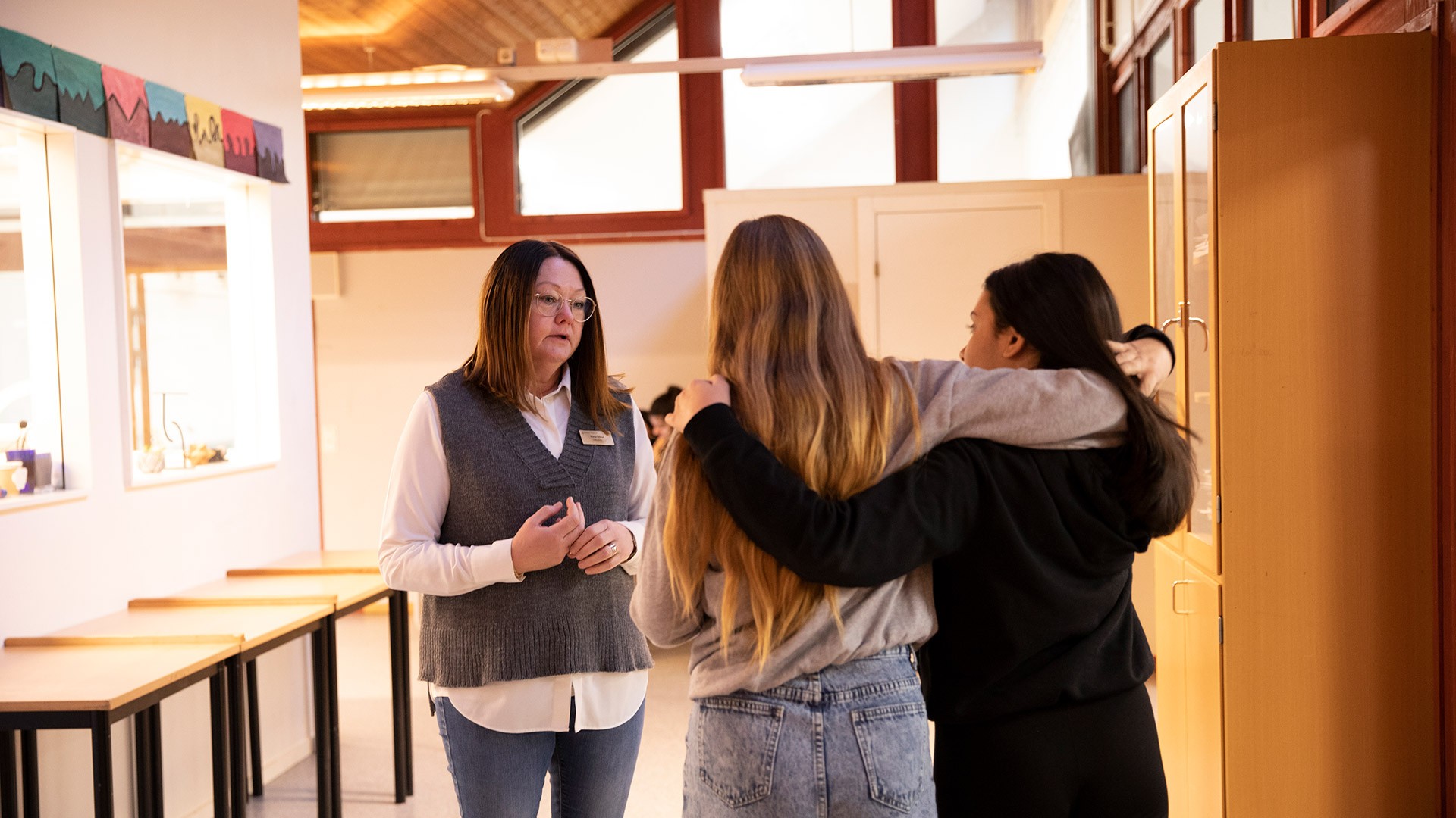 Bilden är tagen inomhus i en lokal på skolan och visar ryggtavlorna på två elever som står med armarna om varandra och som pratar med en kvinna som arbetar på skolan. 