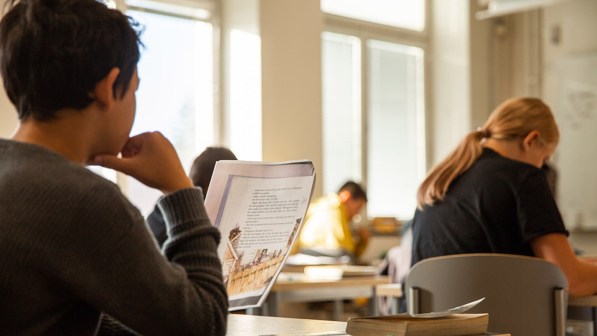 Bilden visar ett klassrum och ryggtavlan på en pojke som läser ett papper. Framför honom syns flera andra elever som ägnar sig åt skolarbete. 