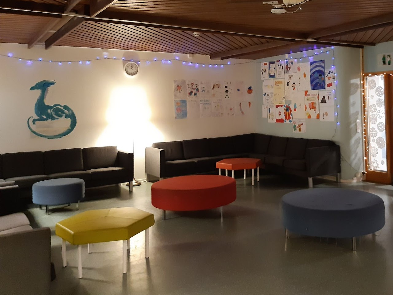 Bilden visar ett uppehållsrum med dämpad belysning, ljusslinga på väggen, soffor och stora sittpuffar. 