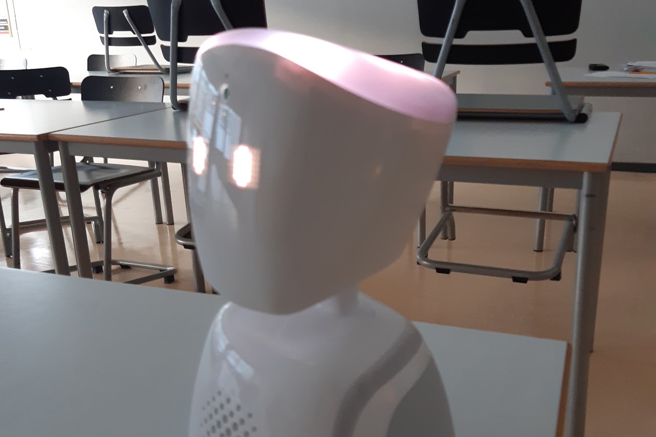 Bilden visar en ljusgrå robot med lysande ögon i ett klassrum med bänkar och stolar. 