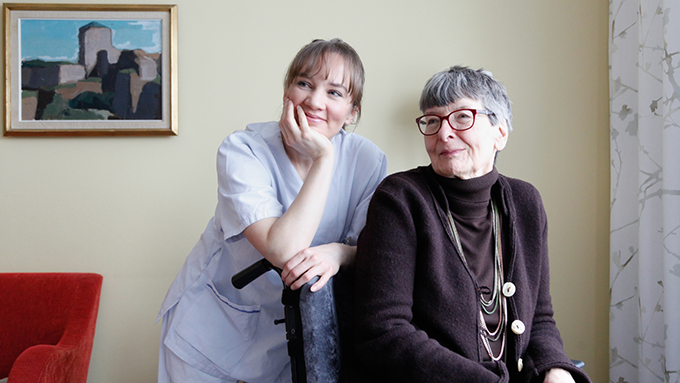 Bild på en äldre leende kvinna i rullstol. Bredvid henne står en ung kvinna som arbetar inom äldreomsorgen.