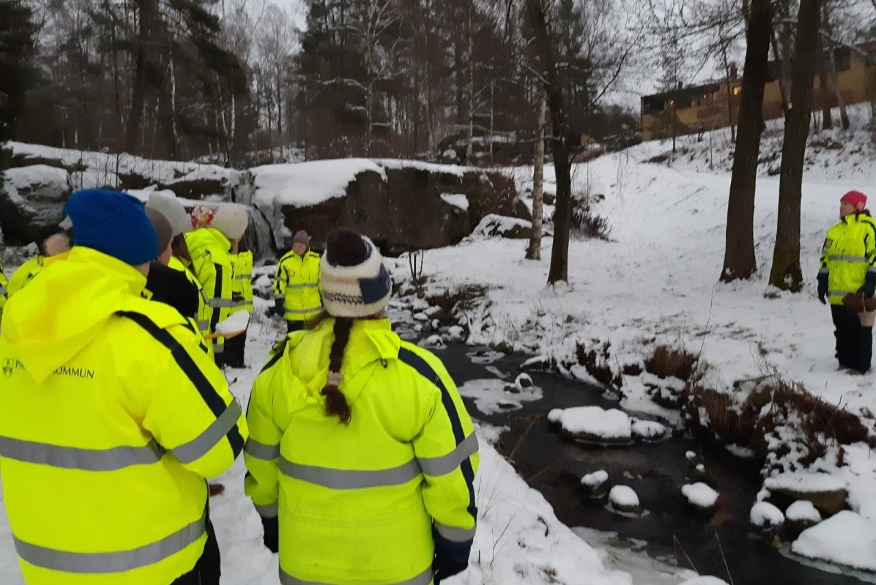 Bilden visar flera personer som står iklädda gula varseljackor i ett vintrigt landskap intill en bäck. 