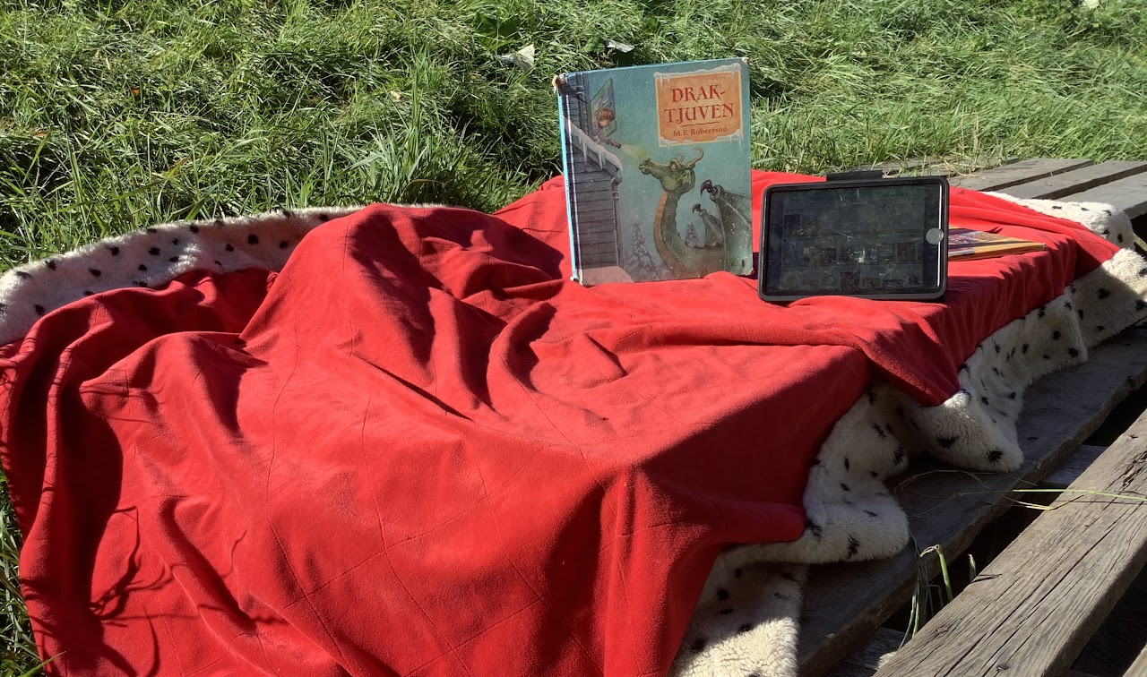 Bilden visar två stycken EU pallar med en kungamantel utbredd, på den står en bok med titeln Draktjuven och bredvid boken ligger en läsplatta. 